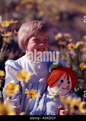 Beautiful young girl holding a Raggety Ann doll and standing in a field of yellow flowers Stock Photo