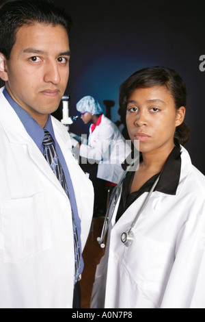 Pair of healthcare professionals look into camera while support team continues research in background. Stock Photo