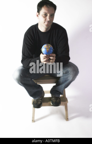 Man in casual clothes sat down on a stool holding a globe in a confident way staring at the camera Stock Photo