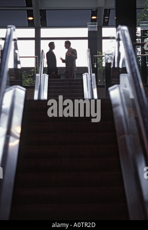 Silhouette of two male executives animated and talking and gesturing at the top of a long flight of escalators in the convention center Stock Photo