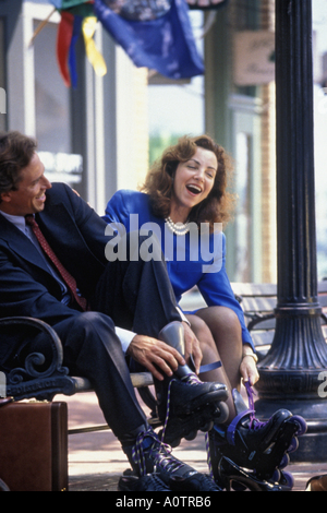 Caucasian businesswoman and businessman taking a break at lunch, sitting on a bench, laughing and putting on roller blades Stock Photo