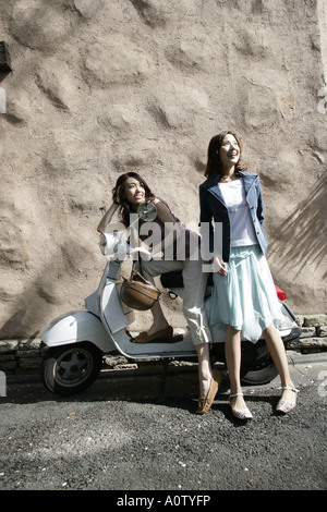Side profile of a young woman sitting on a scooter with her friend standing beside her Stock Photo