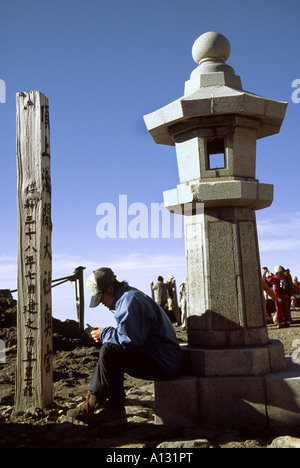 A climber writes a post card at the summit of Mount Fuji while sitting on a large stone lantern called an ishidourou. Japan Stock Photo