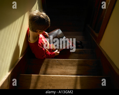 five year old boy in red pj sitting on wood stairs quietly reading comic book with warm morning light streaming in through slatt Stock Photo