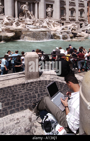 Historical Trevi Fountain statues & sculptures man sitting on paving using laptop computer with camera in hand presumably to upload photo Rome Italy Stock Photo