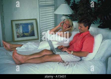 A man and woman sitting in bed The woman is listening to music with headphones and the man is working on a laptop computer Stock Photo