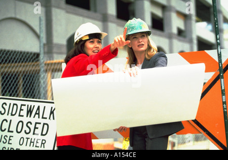 Two female architects holding blue prints and wearing hardhats while inspecting a work site Stock Photo