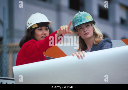 Two female architects holding blue prints and wearing hardhats while inspecting a work site Stock Photo