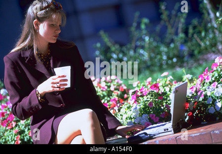 A female executive sitting on a brick wall with flowers blooming in the background holding a drink and working on her laptop Stock Photo