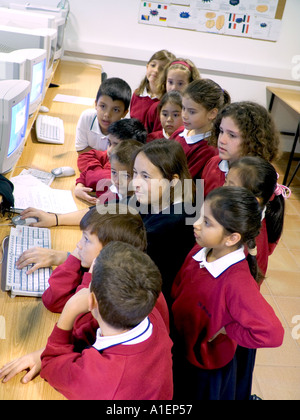 COMPUTER CLASS Infant and junior school children in uniform group around female teacher who is using computer keyboard in school computer classroom Stock Photo