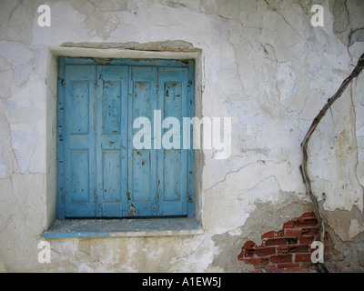 Blue shuttered window in damaged white wall with crack exposing red bricks Stock Photo