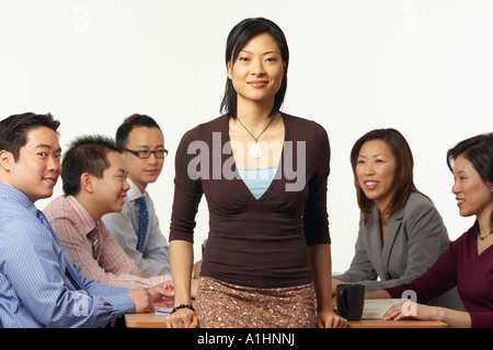 Portrait of a businesswoman leaning against a table with business executives sitting behind her Stock Photo