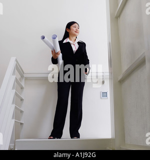 Low angle view of a female architect standing on the staircase and holding rolled up blueprints Stock Photo