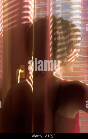 Young woman standing behind curtain looking through window blinds Stock Photo