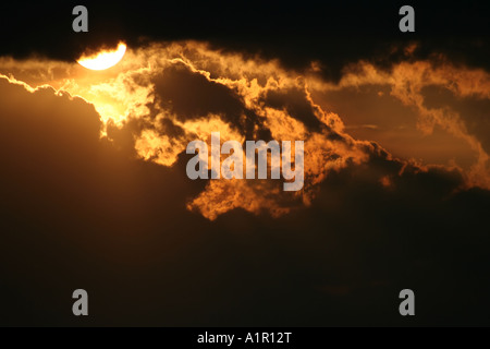 Dramatic sunset with the sun partially hidden behind dark clouds, showcasing the beauty and intensity of a fiery evening sky. Stock Photo