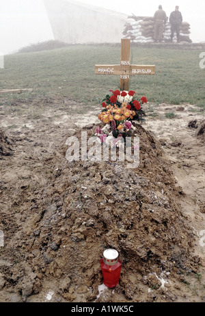WOODEN CROSS AND FRESH FLOWERS ON NEW GRAVE DATED 1955-1991  WHILST SOLDIERS STAND GUARD AT SANDBAGGED MACHINE GUN POSITION Stock Photo
