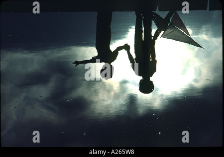 Silhouette of two brothers preparing to launch their homemade wooden schooner in a suburban neighborhood park's glassy blue pond Stock Photo