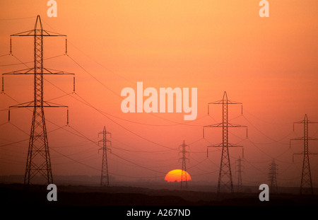 A dramatic orange and red silhouette of  massive electric power lines and towers in a field  overlooking a city at dusk. Stock Photo