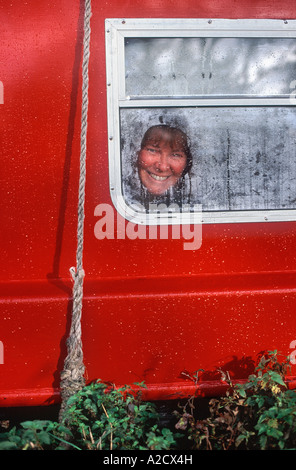 Smiling face of woman looking through cleared dew on window of red narrowboat, River Thames near Abingdon, Oxfordshire, England Stock Photo