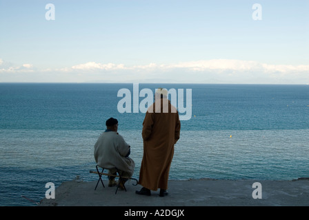 Men in traditional dress looking out to sea across the strait of Gibraltar, Tangier, Morocco Stock Photo
