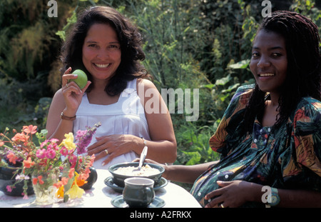 portrait Black pregnant woman enjoying brunch with Hispanic pregnant woman in garden Stock Photo