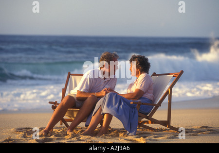 Mature couple holding hands and gazing into each others eyes as they sit in folding chairs on the beach Stock Photo