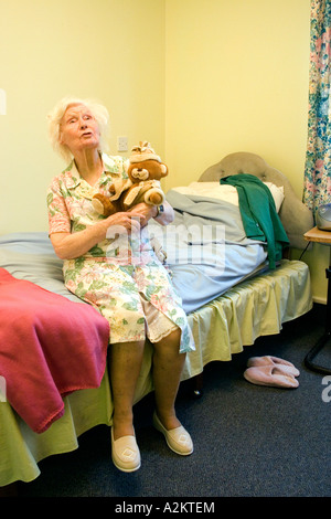 sad old lady in nursing home with senile dementia sitting on edge of bed holding teddy bear