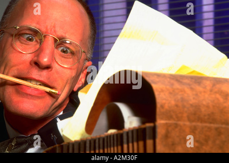 Stressed businessman wearing glasses trying to talk on the phone holding a pencil in his teeth and looking through paperwork Stock Photo