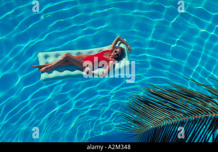 A young woman in a red bathing suit holding a drink while floating on a raft in the crystal blue water of a swimming pool Stock Photo