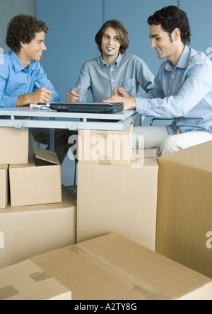 Three male colleagues working at table, cardboard boxes in foreground Stock Photo
