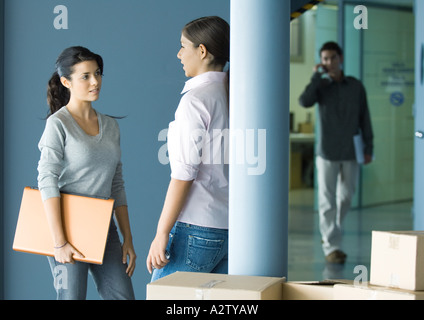 Two young women standing face to face talking, man in background using cell phone Stock Photo