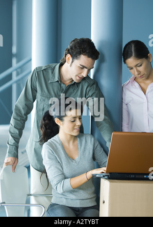 Woman sitting, using laptop on cardboard box, two colleagues looking over her shoulder Stock Photo
