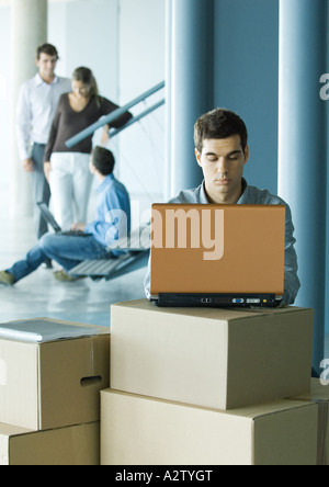 Man using laptop on stack of cardboard boxes, colleagues talking in background Stock Photo