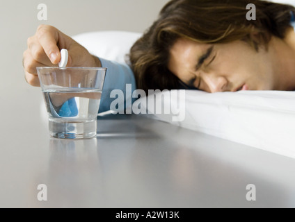 Man lying in bed, squinting eyes shut and holding effervescent medicine over glass of water Stock Photo