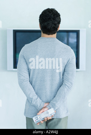 Man standing in front of widescreen TV, holding two remote controls, rear view Stock Photo