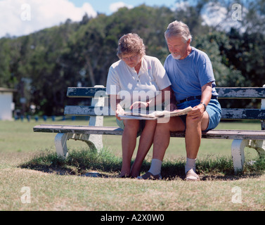 Older couple studying a newspaper whilst seated on park bench. Stock Photo