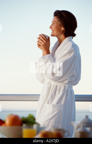 Young woman standing on waterside terrace, wearing white robe and sipping a drink. Stock Photo