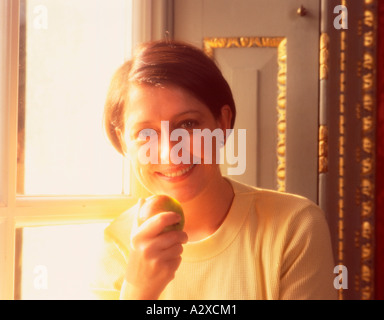 Indoor portrait of natural looking smiling young woman eating apple. Side lit by sunlight from window. Stock Photo