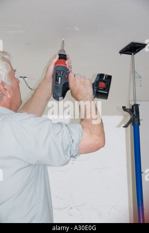 Man fixing plasterboard ceilings using battery power cordless electric drill with screw driver bit & adjustable props to keep sheets in place UK Stock Photo