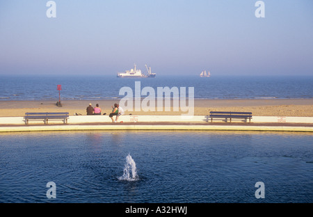 View across model boating lake and wall with two obese couples to beach with blue sky and sea with merchant ship and large yacht Stock Photo