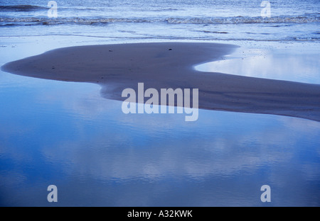 Small waves in a blue-grey sea breaking onto wet sandy beach with only a small dry island of sand and cloud reflections Stock Photo