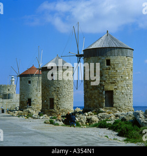 Greek lady in black sitting on rocks with man taking her photograph in front of three windmills at Mandraki Harbor Rhodes Greece Stock Photo