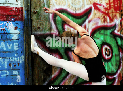 ballet dancer with leg up and arm extended  over head in a stretching pose with graffiti filled wall behind her Stock Photo