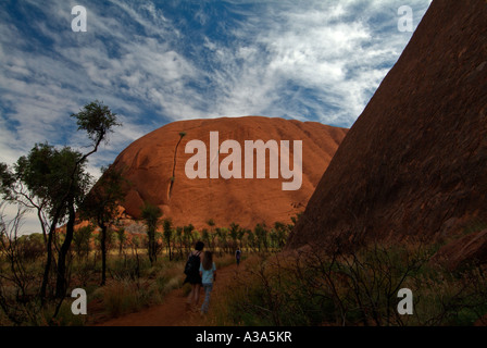 Two tourists walking hiking Ayers rock just after sunrise along the base walk around Uluru with scrub in the foreground Stock Photo