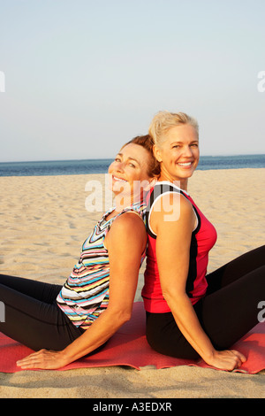 Portrait of two mature women sitting back to back on the beach and smiling Stock Photo
