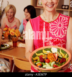 Portrait of a mature woman holding a bowl of salad and her friends sitting at the dining table in the background Stock Photo