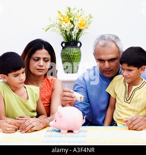 Mid adult man putting an Indian currency note into a piggybank with his wife and two sons beside him Stock Photo