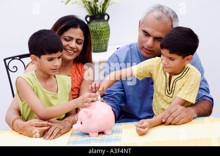 Two boys inserting Indian currency notes into a piggy bank with their parents sitting beside them Stock Photo