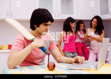 Young man holding a cricket bat and using a laptop in the kitchen Stock Photo
