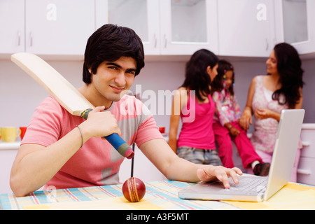 Young man holding a cricket bat and using a laptop in the kitchen Stock Photo